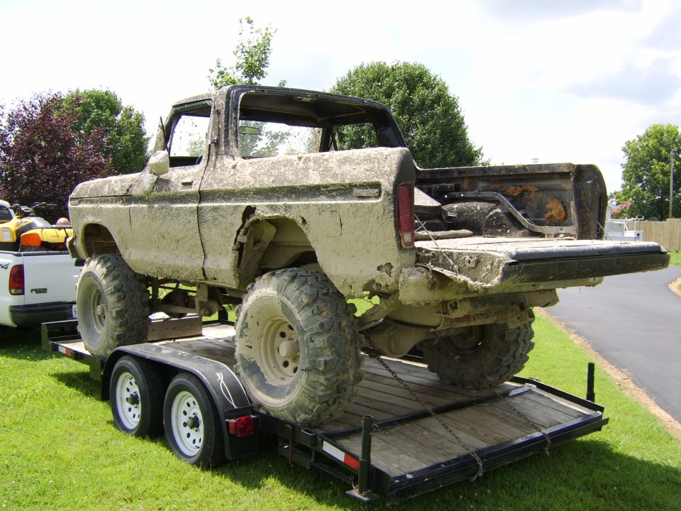 Mud-caked lifted Ford pickup with no doors strapped onto a tandem-axle equipment trailer on grass