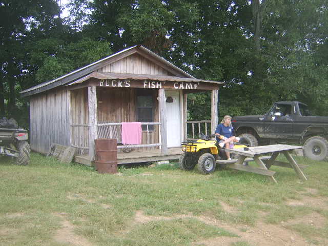 Wooden Buck's Fish Camp cabin with dark lifted Ford pickup, yellow ATV, and person at picnic table