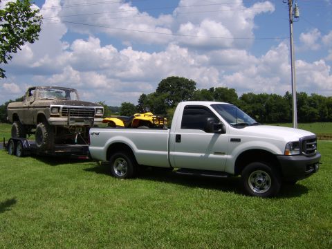 2003 Ford F-250 - A white 2003 Ford F-250 tows a trailer carrying a lifted truck and an ATV on a grassy field under a cloudy sky.