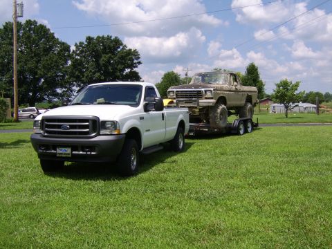 2003 Ford F-250 - White Ford Super Duty tows a lifted, older Ford pickup truck on a trailer across a grassy field.