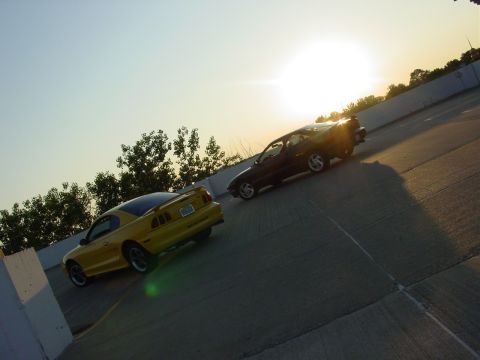 1998 Ford Mustang - Two cars drive on a highway during sunset, with a yellow Ford Mustang in the foreground.