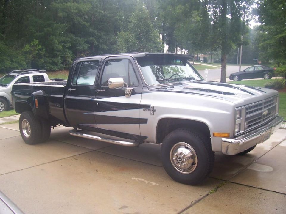 Dually pickup truck, two-tone black and silver with racing stripes, parked on a driveway. Another white SUV is partially visible to the left