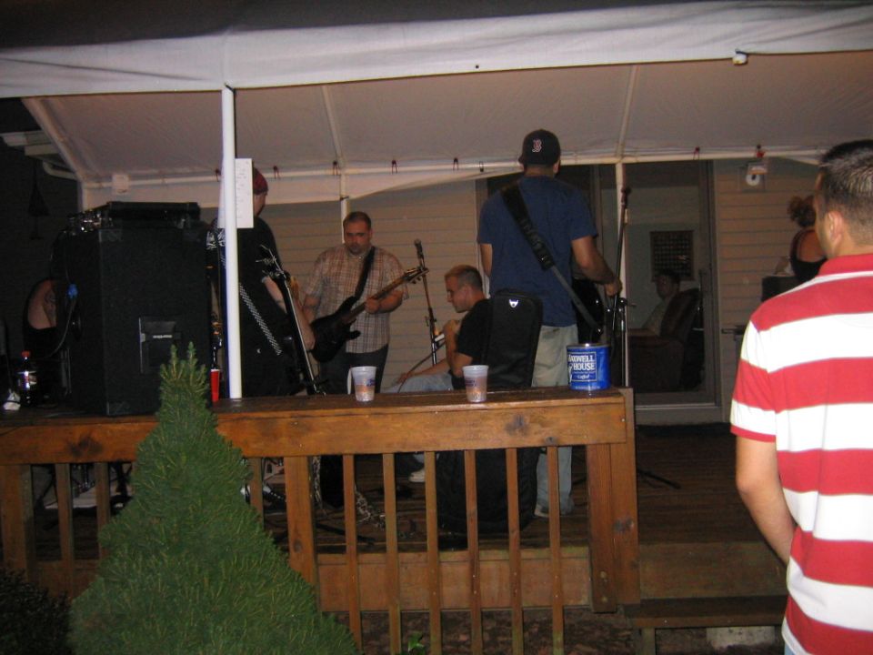 Band performs on a wooden deck at night, with speakers and instruments visible