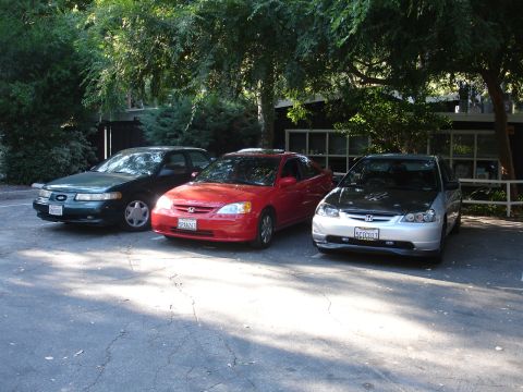1995 Ford Taurus - Three cars are parked in a lot under trees. The vehicles include a green sedan, a red coupe, and a silver hatchback.