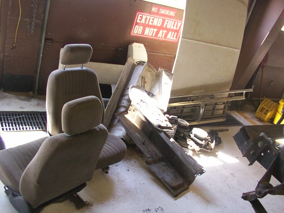 Two car seats and various vehicle parts are piled on a garage floor. A sign reads 'NO SMOKING EXTEND FULLY OR NOT AT ALL'
