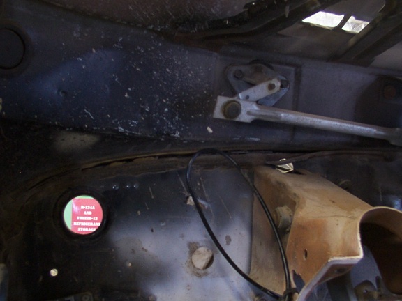 Underside of a vehicle showing the wiper linkage and a red circular sticker with text