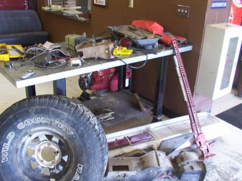 1978 Ford Bronco - A workbench is covered in automotive parts and tools, with a large tire and a red Hi-Lift jack also visible in the scene.