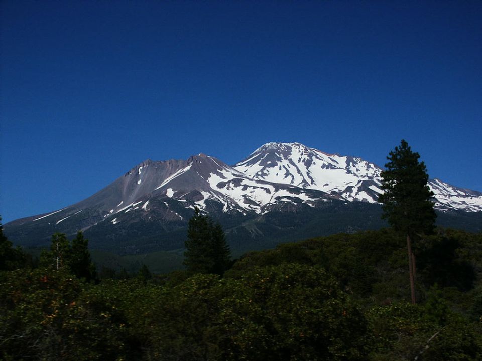 Snow-capped mountains under a clear blue sky, with dark green trees in the foreground. A watermark reads SUPERMOTORS.NET