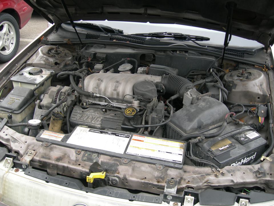 engine bay with V6 engine, battery, and some hoses. Another car's wheel is visible on the left
