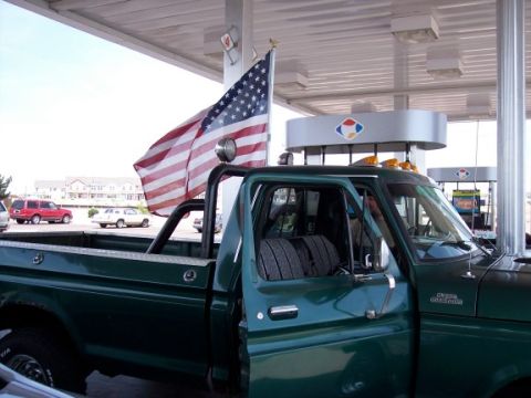 1972 Ford Ranchero - A green Ford pickup truck flies an American flag while parked at a gas station.