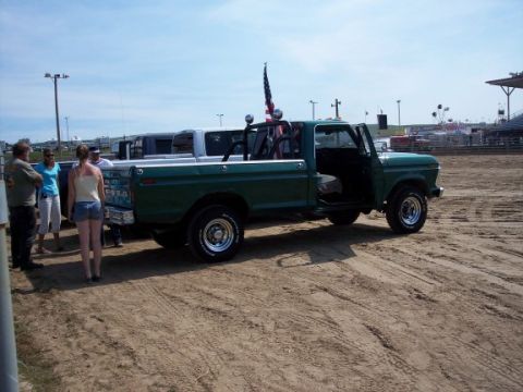 1972 Ford Ranchero - A green pickup truck flies an American flag from its roll bar. The truck is parked on a dirt surface with other vehicles and spectators in…