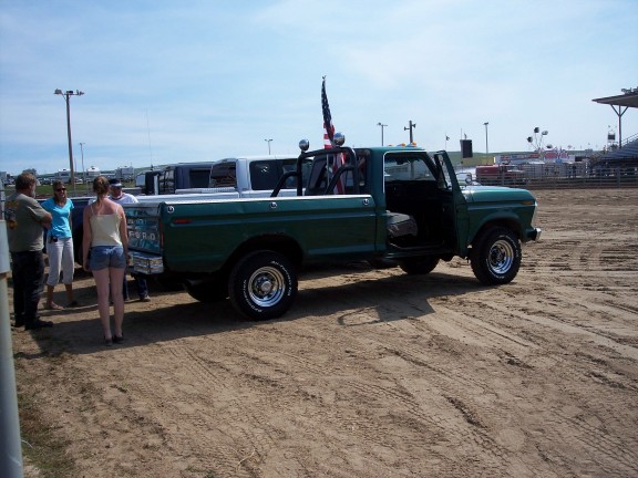 Green 1970s Ford long-bed pickup with roll bar lights and flag parked on a dirt fairgrounds arena