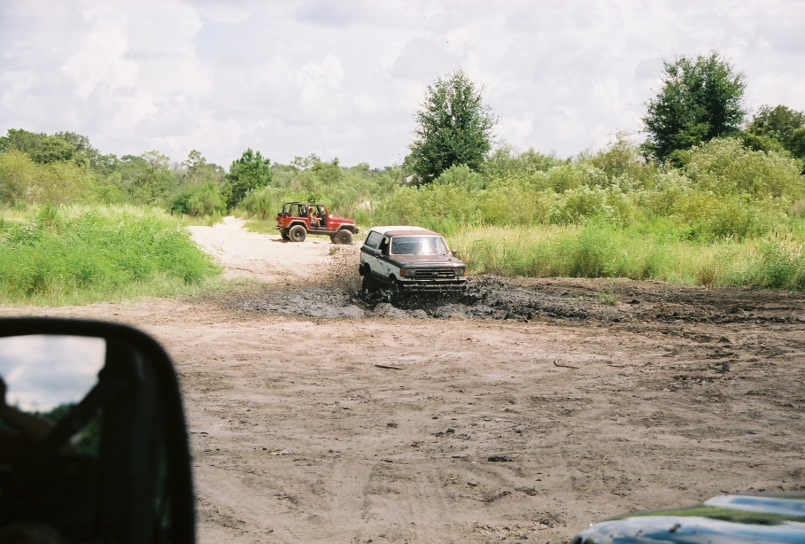 Brown and white Ford Bronco stuck in mud, front view. Another red Jeep is visible in the background