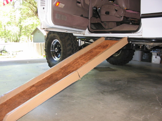 White lifted Ford Bronco on off-road tires with a carpeted ramp leaning up to its underside in a garage