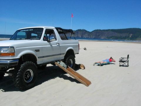 1995 Ford Bronco - A white 1995 Ford Bronco is parked on a sandy beach with a person relaxing in a lounge chair next to it.