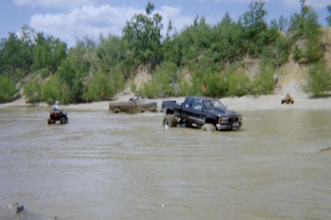 2005 Dodge 1500 - Multiple trucks and ATVs navigate through a muddy water area with trees lining the banks.