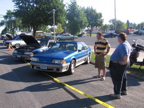 1989 Ford Mustang - Two men observe a blue Ford Mustang hatchback parked at an outdoor event with other vehicles present.