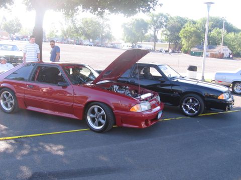 1989 Ford Mustang - Two Ford Mustangs, a red one with its hood up, are parked side-by-side at an outdoor event.