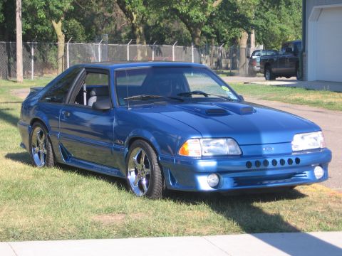 1989 Ford Mustang - This blue 1989 Ford Mustang is parked on a grassy area next to a building.