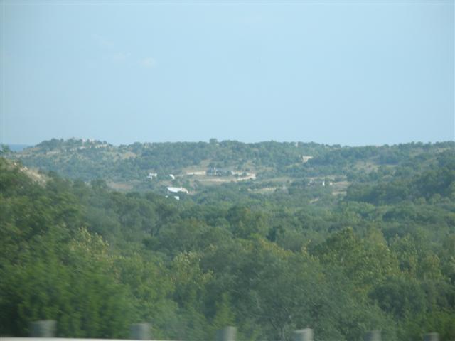 Hazy hill-country landscape of wooded ridges and scattered buildings shot from a moving vehicle