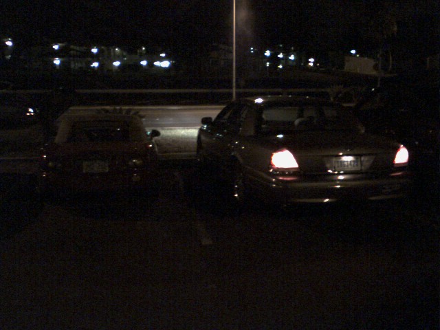 Two sedans parked at night in a dark lot, one with glowing red taillights and lit license plate