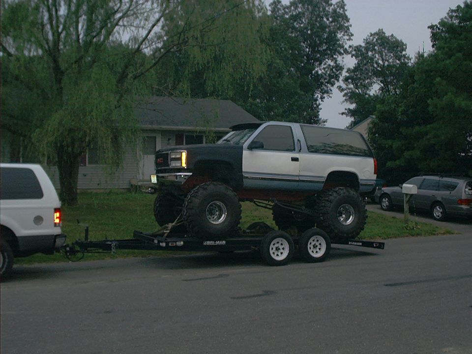 Lifted GMC SUV on a trailer being towed by a white SUV. Another car is parked in the background