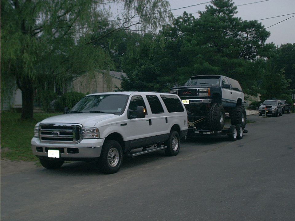 White Ford Excursion towing a trailer loaded with a lifted black GMC SUV on mud tires