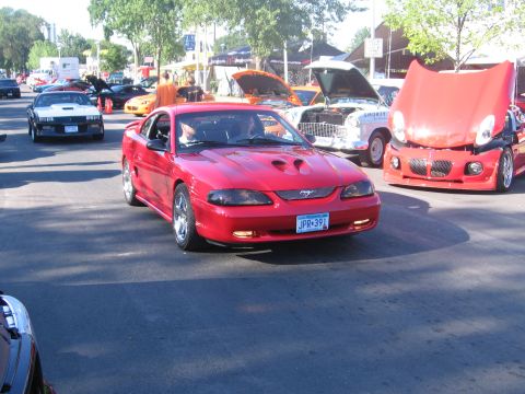 1989 Ford Mustang - A red Ford Mustang drives down a street lined with trees and other vehicles.