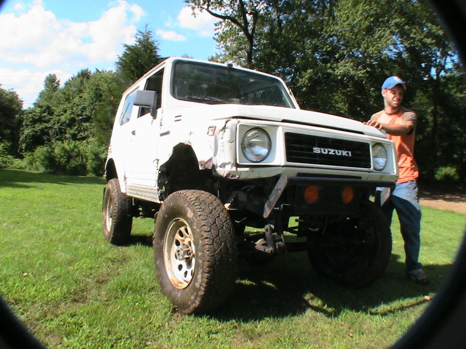 Lifted white Suzuki SUV with big off-road tires on grass, man in orange shirt standing beside it