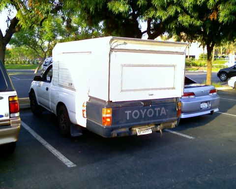 2004 GMC Sierra 1500 - A white Toyota pickup truck is parked with a white camper shell covering the bed. Another car is parked to its right.