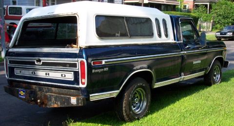 1975 Ford F-150 - A dark blue Ford pickup truck with a white camper shell is parked on a grassy area next to a road.