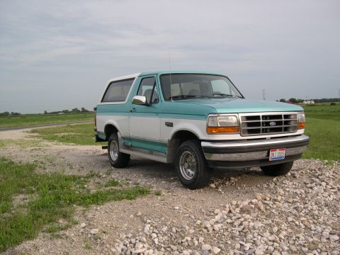 1994 Ford Bronco - A two-tone teal and white Ford Bronco is parked on a gravel lot with a field and power lines in the background.