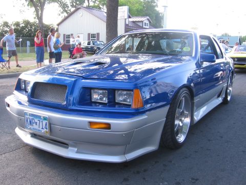 1989 Ford Mustang - A custom blue-and-silver 1980s Ford Thunderbird coupe parked at an evening cruise-in, shown from the low front three-quarter with a hood sc…