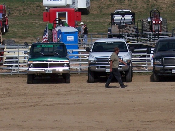 Green vintage Ford pickup and white Dodge Ram parked in a dirt truck-pull arena pit area