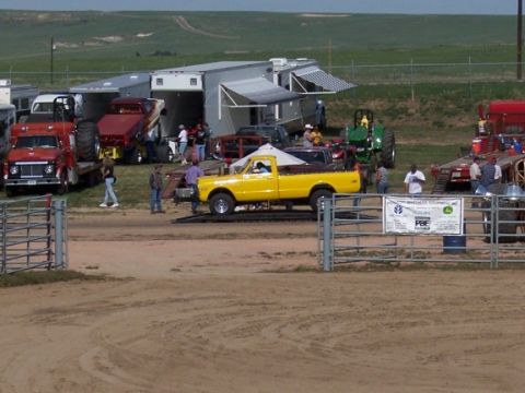 1972 Ford Ranchero - A yellow pickup truck is positioned on a ramp, with a trailer and tractor visible in the background at an outdoor event.