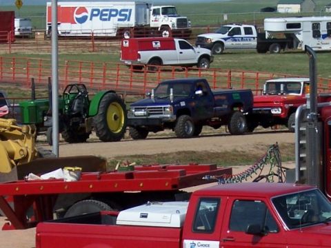 1972 Ford Ranchero - A blue pickup truck is seen towing a red trailer, with various other vehicles and farm equipment in the background.