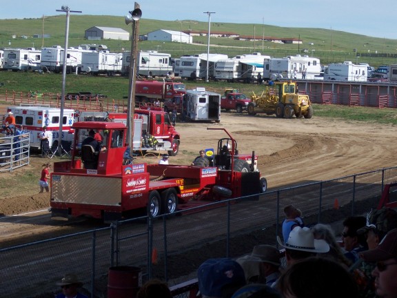 Red flatbed truck moving a weight-transfer sled on a dirt pulling track at a fairground