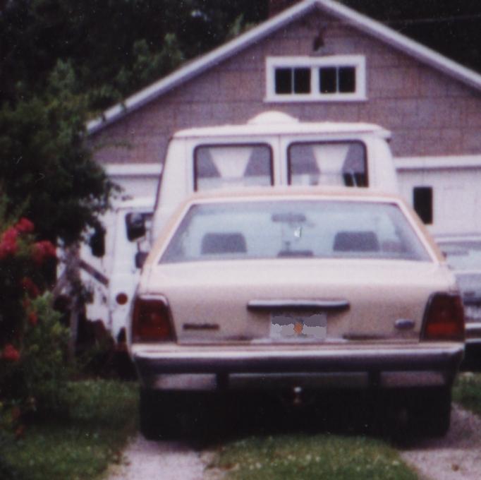 Rear view of a tan Ford sedan in a driveway behind a white van, house in background