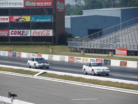 1992 Ford Taurus - Two Ford Tauruses compete on a drag strip, with grandstands and sponsor banners in the background.