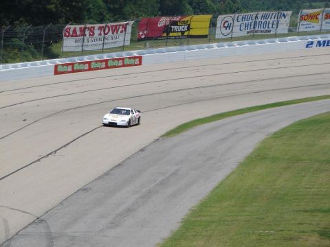 1992 Ford Taurus - A white race car navigates a turn on a paved racetrack with various sponsor banners in the background.