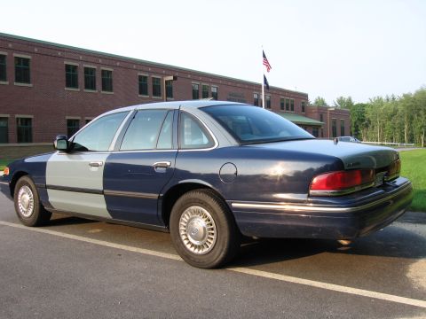1991 Ford LTD Crown Victoria - A two-tone Ford Crown Victoria sedan is parked in front of a brick building with an American flag flying.