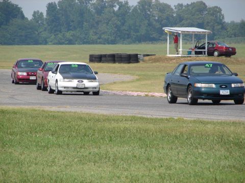 1992 Ford Taurus - Cars are lined up on a race track, with a pit stop area in the background.