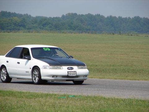 1992 Ford Taurus - A white 1992 Ford Taurus with a black hood is seen on a race track with a driver inside.