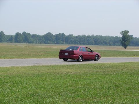 1992 Ford Taurus - A red 1992 Ford Taurus sedan is seen from the rear on a paved track with grassy fields and trees in the background.