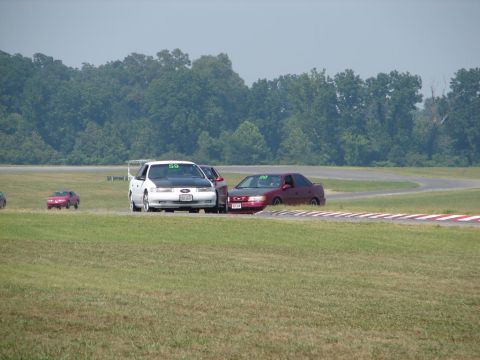 1992 Ford Taurus - A white 1992 Ford Taurus with a black hood and a large rear spoiler is on a race track, with other cars nearby.