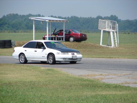 1992 Ford Taurus - A white 1992 Ford Taurus with a black hood drives on a race track, with another red car in the background.