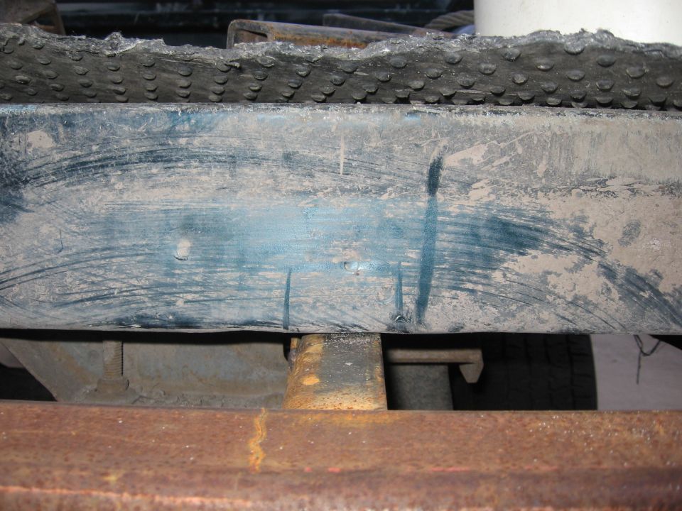 Rusty metal frame with a blue-painted bumper above, showing scuff marks and dirt. A black textured mat is visible at the top