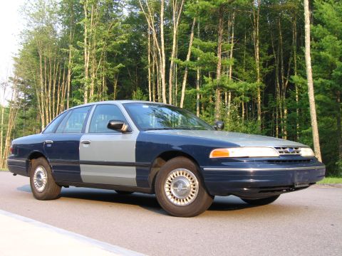 1991 Ford LTD Crown Victoria - A two-tone sedan, blue and gray, is parked on an asphalt road with a forest of trees behind it.