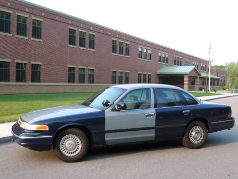 1991 Ford LTD Crown Victoria - A two-tone police car, with blue and grey paint, is parked on a street in front of a large brick building.