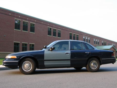 1991 Ford LTD Crown Victoria - Side view of a two-tone police car with dark blue and light gray panels, parked in front of a brick building.